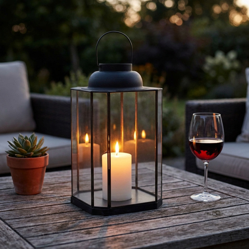 Lanterna decorativa illuminata da una candela su un tavolo da giardino, affiancata da un calice di vino rosso e una piccola pianta succulenta.