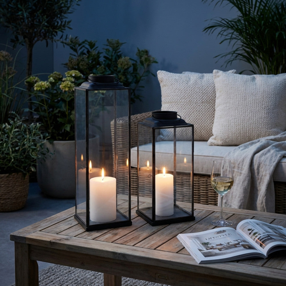 Lanterna Mollie Piccola in acciaio e vetro illuminata da una candela, posizionata su un tavolo da giardino all'imbrunire per un'atmosfera sofisticata.
