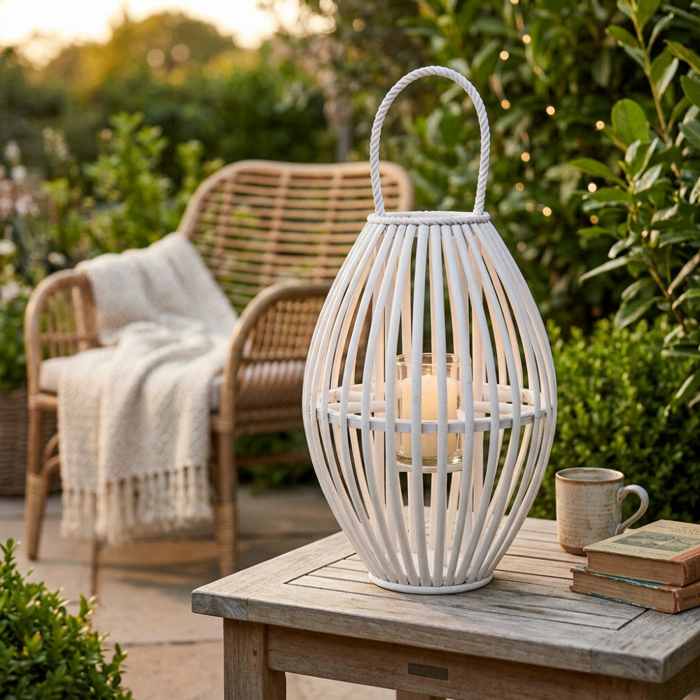Lanterna Seline in vimini bianco posizionata su un tavolino in legno da esterni, con una candela accesa all'interno del cilindro in vetro, circondata da un giardino rigoglioso e una poltrona in rattan.