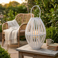 Lanterna Seline in vimini bianco posizionata su un tavolino in legno da esterni, con una candela accesa all'interno del cilindro in vetro, circondata da un giardino rigoglioso e una poltrona in rattan.