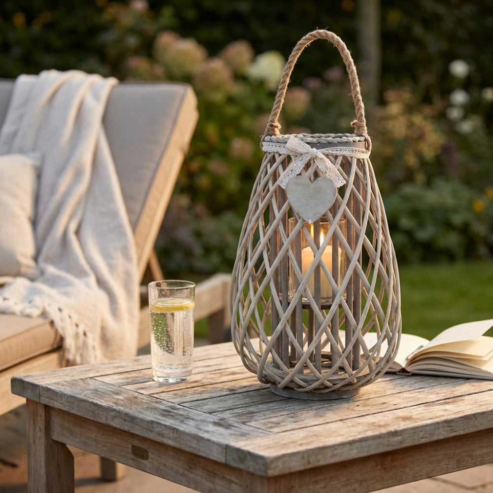 Dettaglio della Lanterna Simonne in vimini intrecciato su un tavolo da giardino in legno grezzo, con un bicchiere d'acqua e un libro, illuminata dalla calda luce serale.