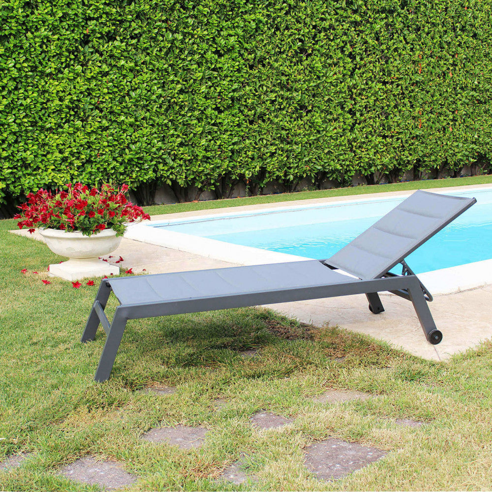 Lettino Formentera in alluminio antracite immerso nel verde di un giardino curato vicino all'acqua.