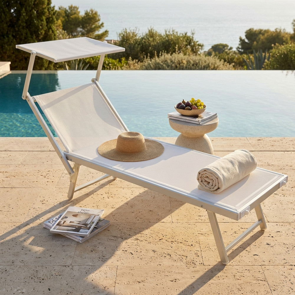 Vista angolare del lettino Jesolo bianco a bordo piscina, decorato con cappello di paglia, telo arrotolato e tavolino in pietra con frutta.