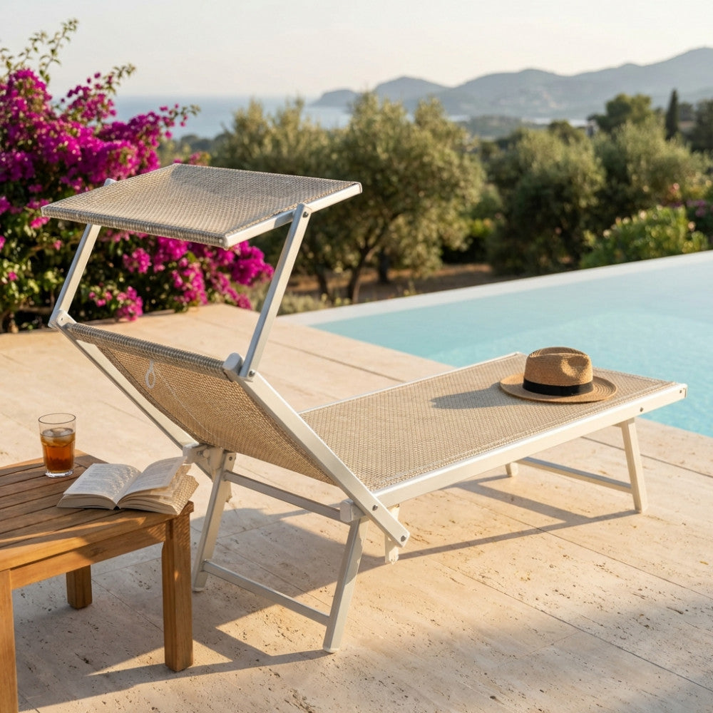 Lettino prendisole Otranto in alluminio e textilene beige a bordo piscina, con cappello di paglia e libro su tavolino in legno.