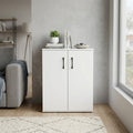 Mobiletto Bennie in legno bianco con due ante, posizionato in un soggiorno moderno e decorato con libri e una pianta.