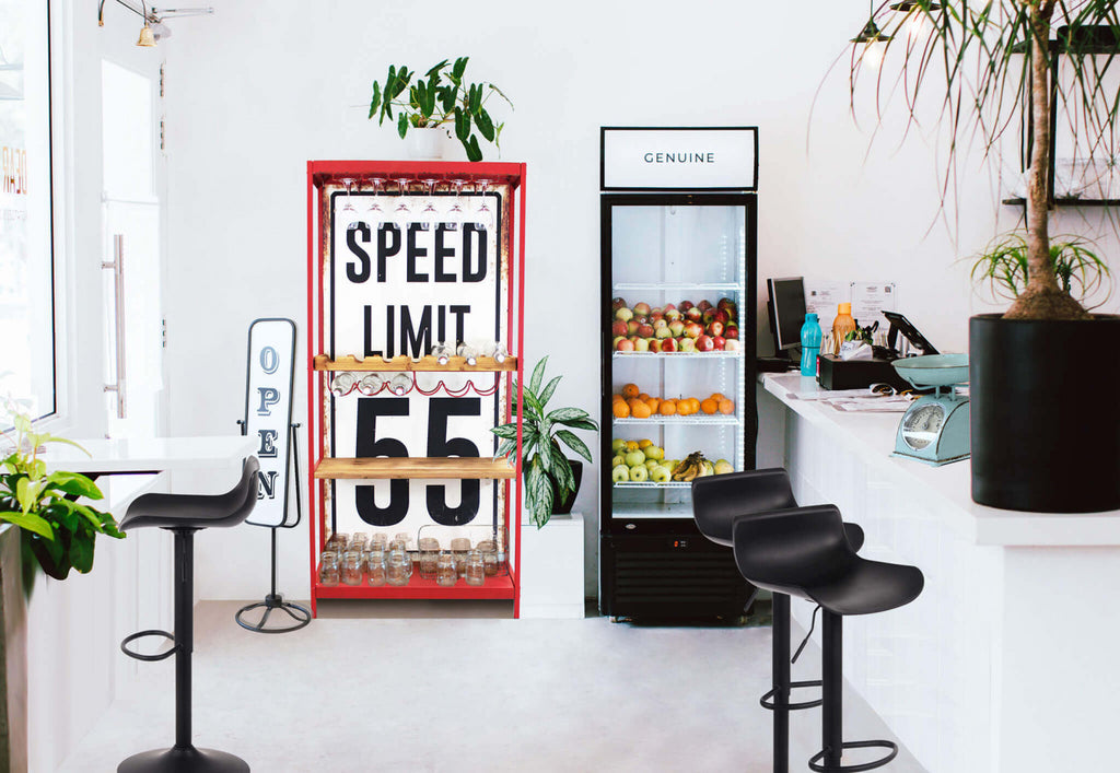 Mobile bar Clayton inserito nell'arredamento di un caffè moderno e luminoso con sgabelli neri e piante.