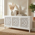 Vista angolata della credenza Jacob in legno di mango intagliato bianco, ambientata in un soggiorno luminoso con vasi decorativi.