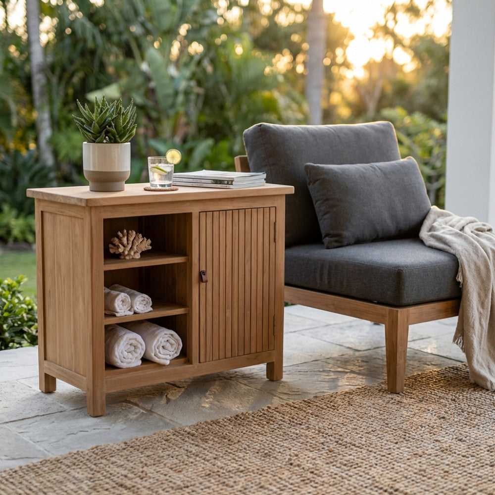 Credenza Lexington 90 in teak premium allestita su un patio in pietra accanto a una poltrona moderna, con asciugamani arrotolati e una pianta in vaso.