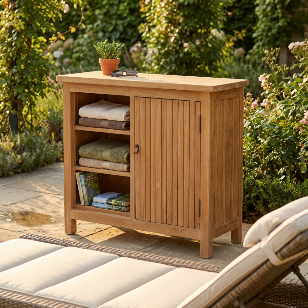 Ambientazione esterna con il mobile in teak Lexington 90 in un giardino rigoglioso, con asciugamani piegati e libri accanto a un lettino intrecciato.