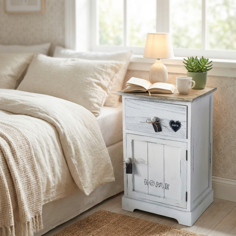 Comodino shabby chic Ander in legno bianco e marrone chiaro, ambientato in una luminosa camera da letto accanto a un letto con lenzuola in lino.