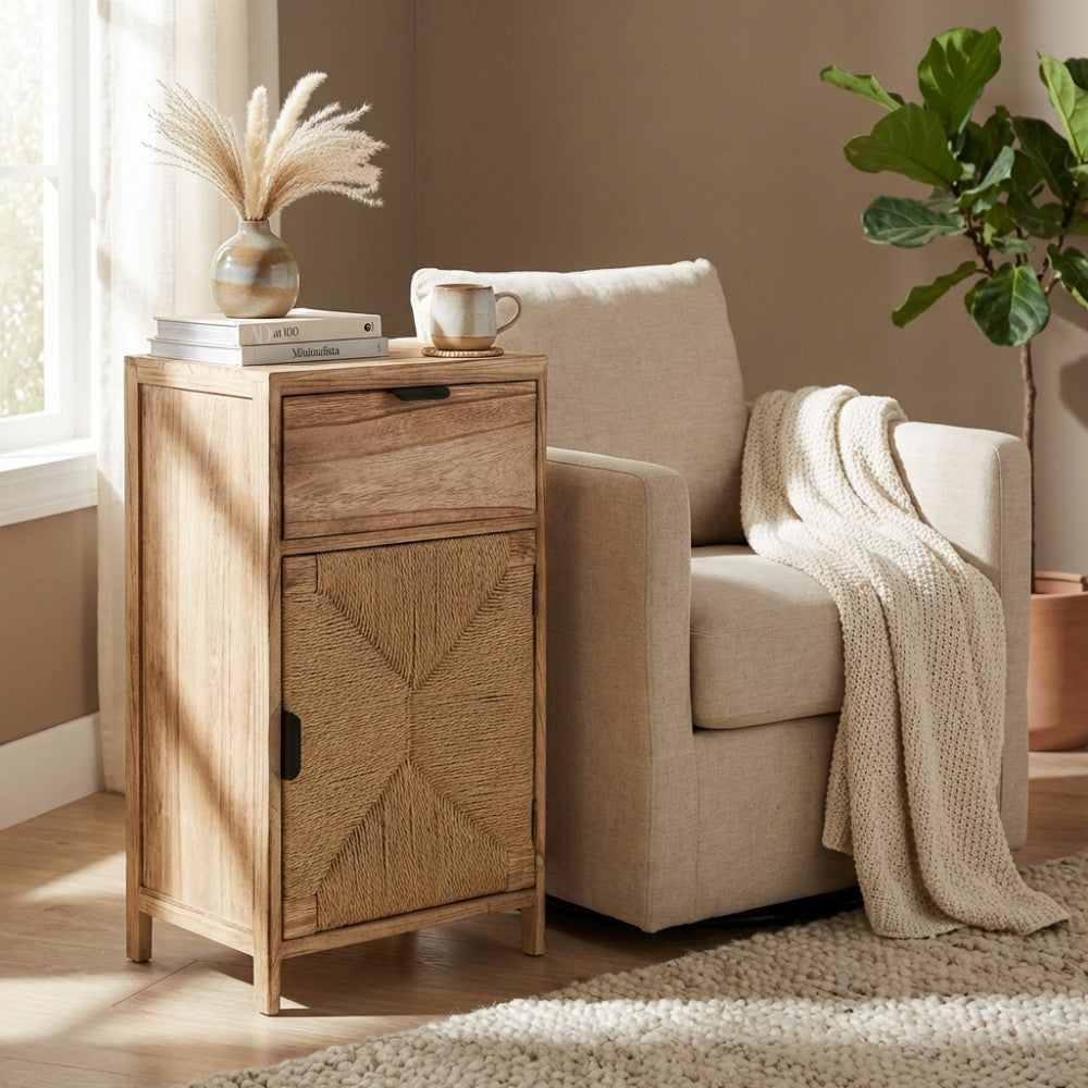 Mobiletto Carlos con anta in corda intrecciata, posizionato come tavolino laterale vicino a una poltrona beige in un salotto dal design naturale.