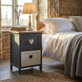 Comodino shabby chic Alvaro in legno grigio e bianco, posizionato in una camera da letto rustica illuminata dalla luce naturale.