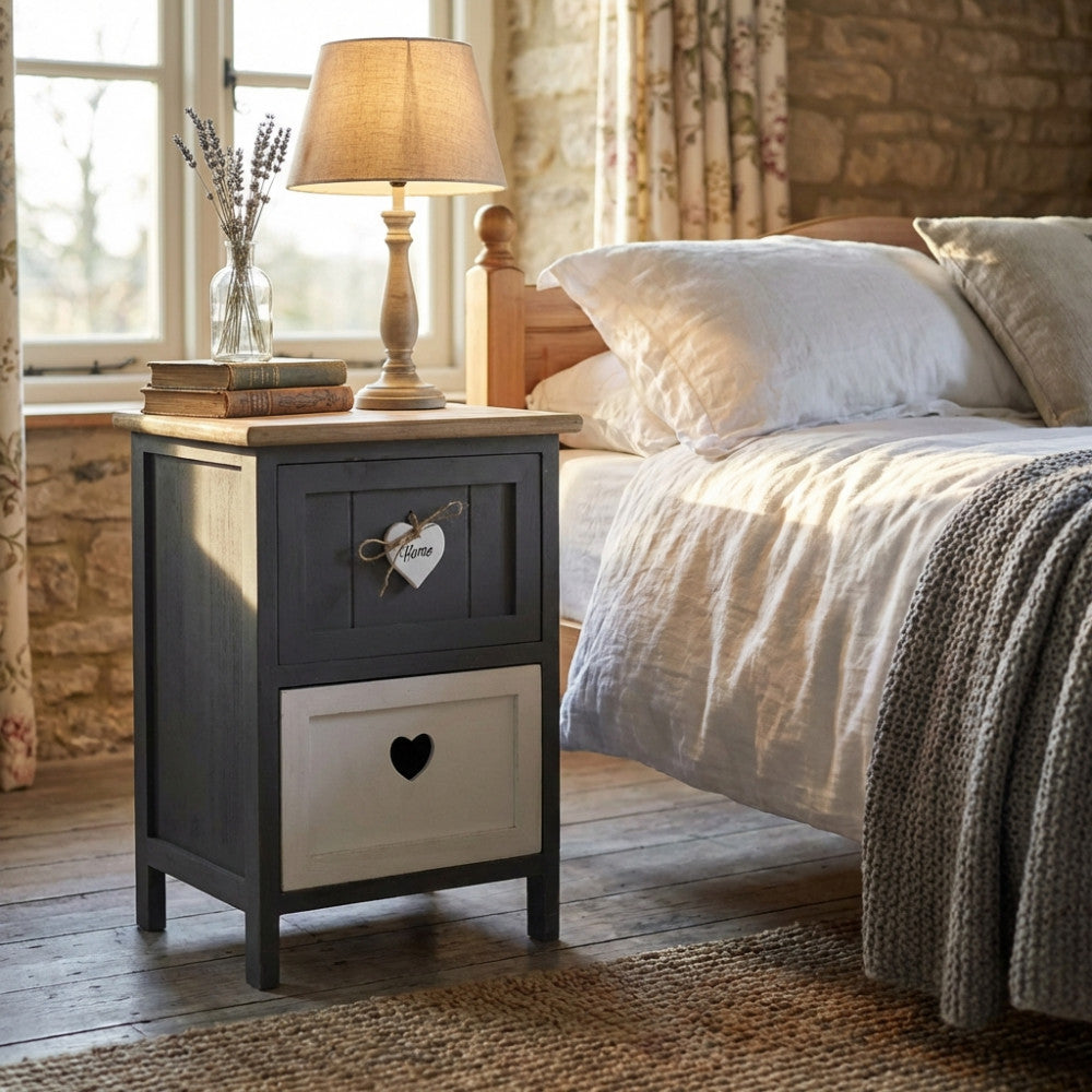Comodino shabby chic Alvaro in legno grigio e bianco, posizionato in una camera da letto rustica illuminata dalla luce naturale.