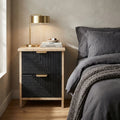 Comodino ASIER in rovere chiaro e legno nero ambientato in una camera da letto elegante con lampada dorata.