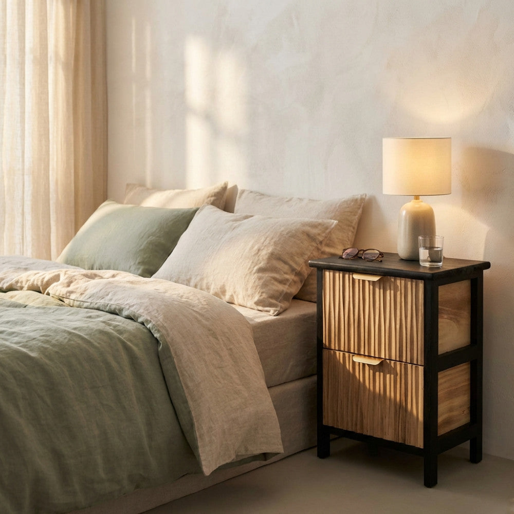 Comodino Cesar moderno in legno scanalato marrone chiaro e struttura nera, posizionato accanto a un letto con lenzuola in lino verde e beige, illuminato dalla luce naturale.