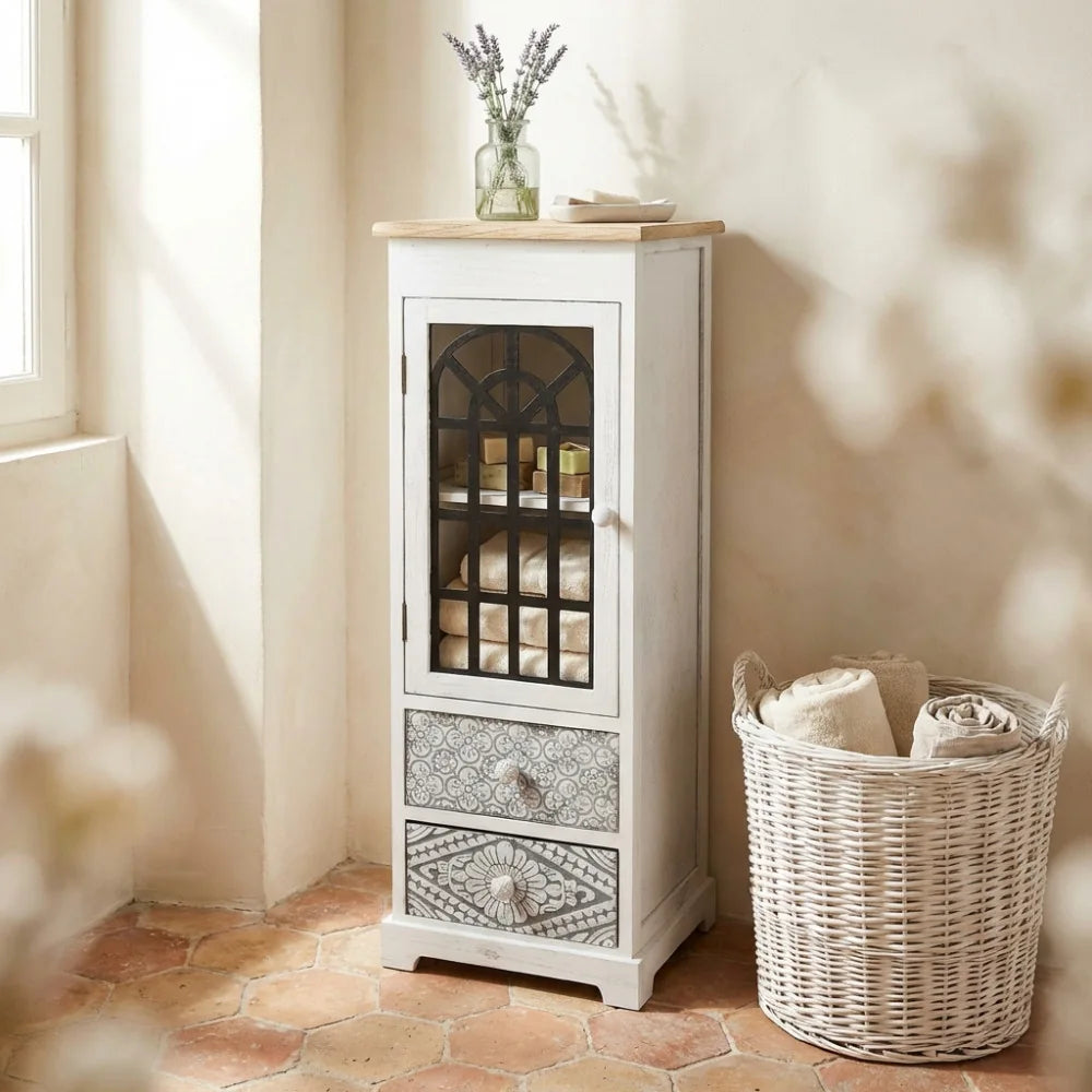 Vetrina shabby chic in legno bianco con anta in vetro e cassetti decorati, ambientata in un bagno luminoso.