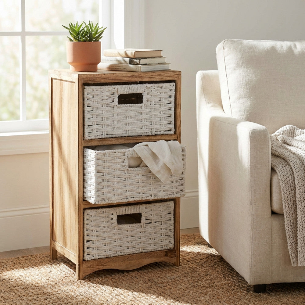 Mobiletto vintage Adrian in legno di paulownia marrone chiaro con tre cassetti a cesto bianco, posizionato accanto a una poltrona beige.