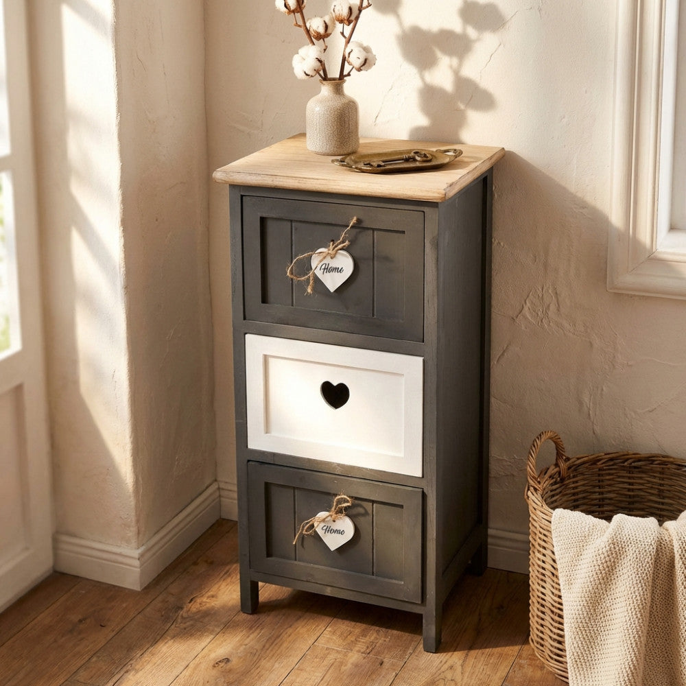 Mobiletto a tre cassetti in stile shabby chic grigio e bianco, posizionato in un angolo luminoso con un vaso di fiori di cotone sulla superficie in legno naturale.