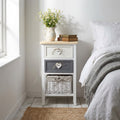 Comodino Andres shabby chic ambientato accanto a un letto con vaso di fiori.