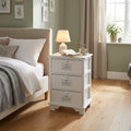 Comodino shabby chic bianco a tre cassetti ambientato di fianco a un letto matrimoniale con lampada accesa.