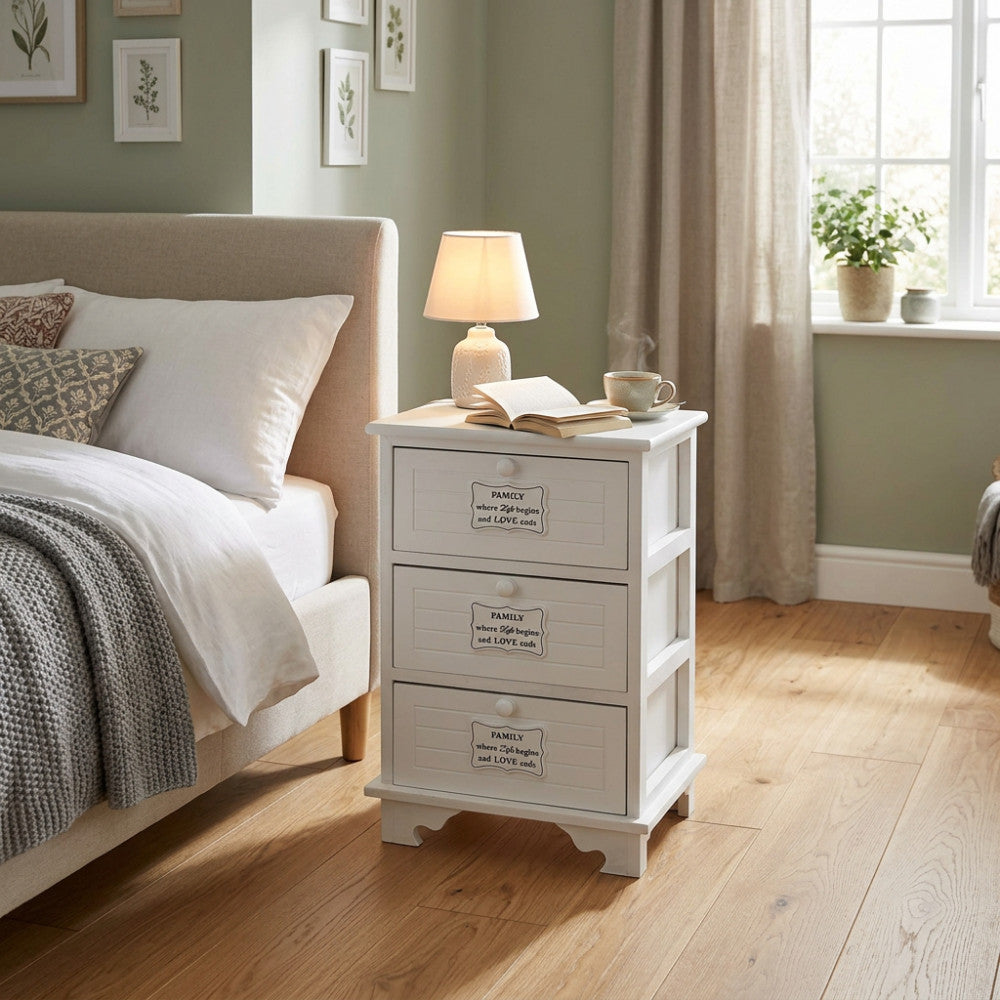 Comodino shabby chic bianco a tre cassetti ambientato di fianco a un letto matrimoniale con lampada accesa.
