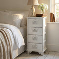 Comodino shabby chic bianco Biel posizionato accanto a un letto con lampada e fiori.