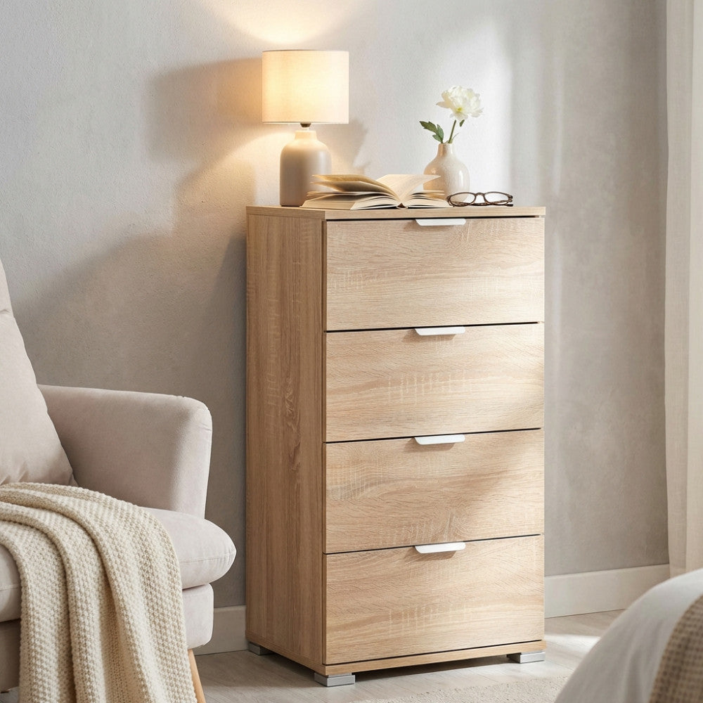 Il mobile a 4 cassetti marrone chiaro posizionato in un accogliente angolo lettura accanto a una poltrona beige, illuminato dalla luce calda di una lampada da tavolo.