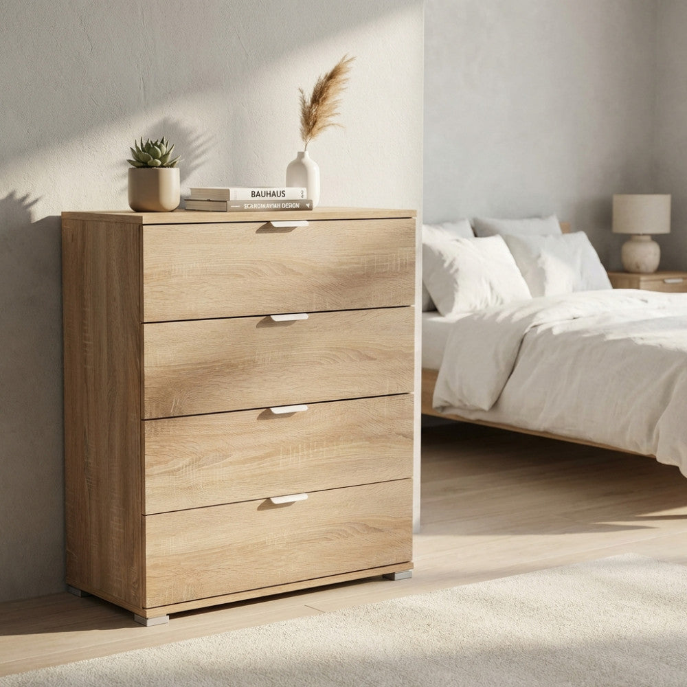 Vista angolare della cassettiera in bilaminato effetto legno, posizionata accanto a un letto matrimoniale con tessuti bianchi e luce naturale.