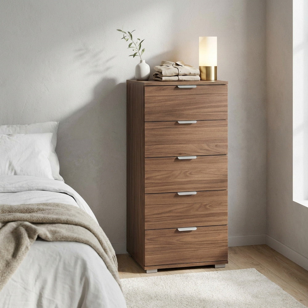 Cassettiera a 5 cassetti in finitura noce posizionata in una camera da letto luminosa, decorata con una lampada moderna e indumenti piegati.