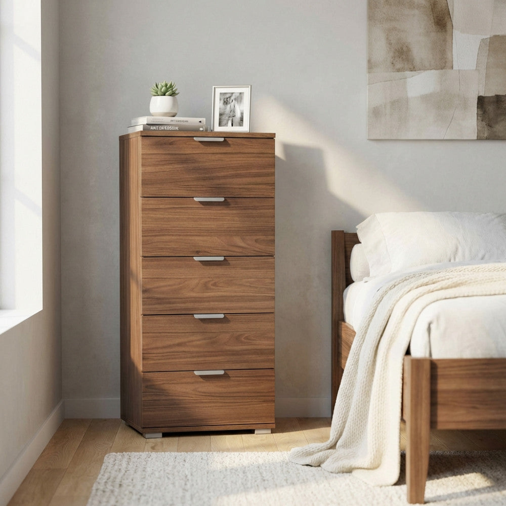 Vista angolata del mobiletto a 5 cassetti color marrone noce, affiancato a un letto in legno con biancheria chiara.