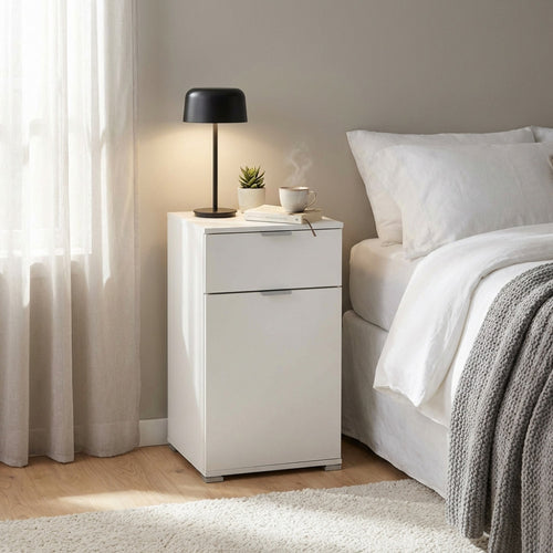 Comodino bianco con cassetto e anta posizionato accanto a un letto, allestito con lampada nera e tazza fumante.