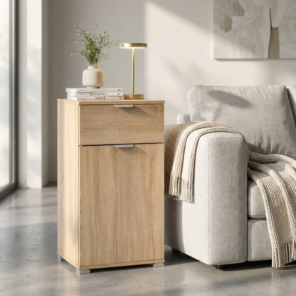 Comodino MR805044 in truciolare bilaminato marrone chiaro, ambientato accanto a un divano grigio con lampada di design.