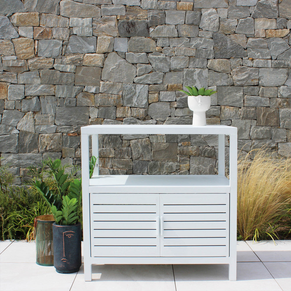Mobiletto Phoenix bianco posizionato contro un muro in pietra naturale, decorato con vasi di design e piante verdi.