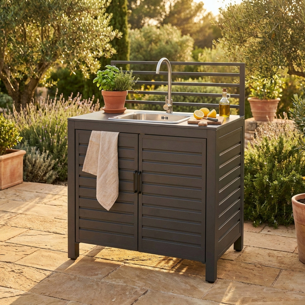 Vista frontale del modulo lavello Oklahoma in alluminio, circondato da piante aromatiche e vasi in terracotta su pavimentazione in pietra.