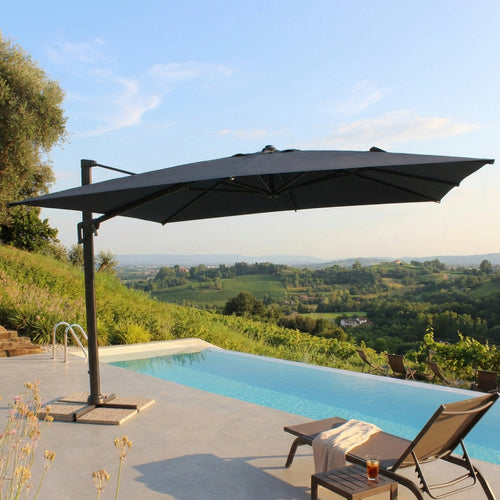 Vista laterale dell'ombrellone Acrux 4x4 che proietta un'ampia ombra su lettini prendisole eleganti in un giardino di lusso.