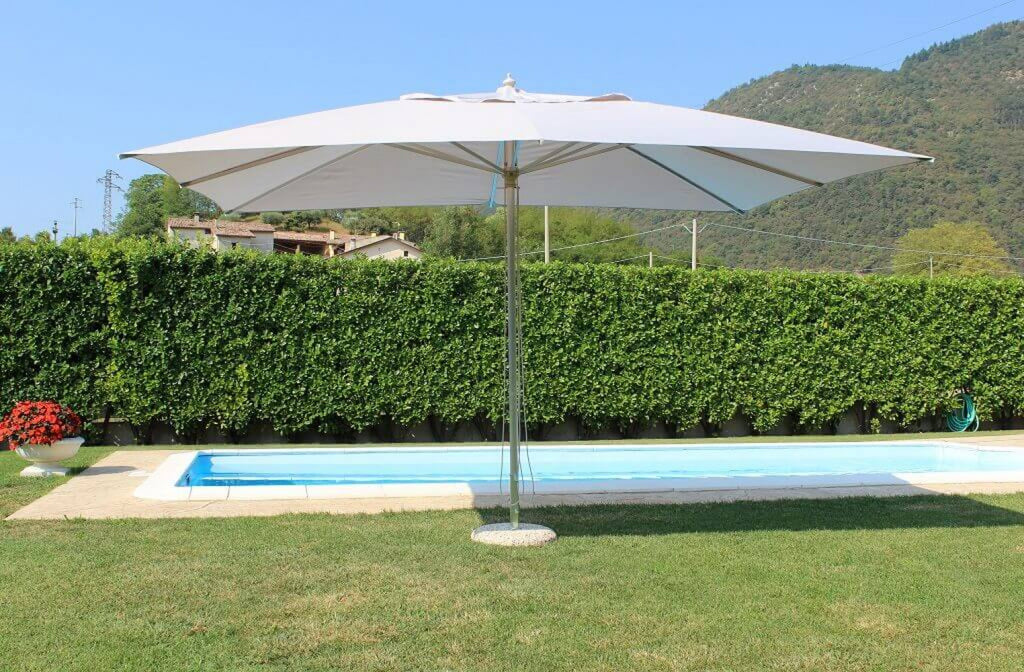 Ombrellone Andromeda posizionato su un prato verde curato vicino a una piscina, evidenziando la sua ampia zona d'ombra.