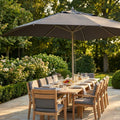 Ombrellone Andromeda grigio aperto su un grande tavolo da pranzo in legno di teak in un rigoglioso giardino illuminato dal sole.
