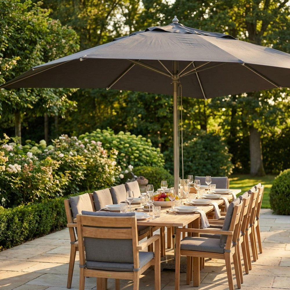 Ombrellone Andromeda grigio aperto su un grande tavolo da pranzo in legno di teak in un rigoglioso giardino illuminato dal sole.