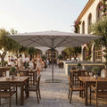 Ombrellone a palo centrale Arneb 5x5 metri in alluminio effetto legno e telo taupe, allestito in un elegante patio di un ristorante all'aperto.