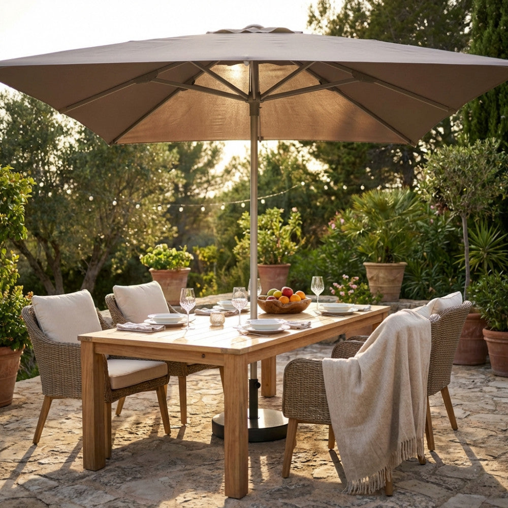 Ombrellone Brian color tortora aperto su un tavolo da pranzo in legno massello in un giardino mediterraneo al tramonto.