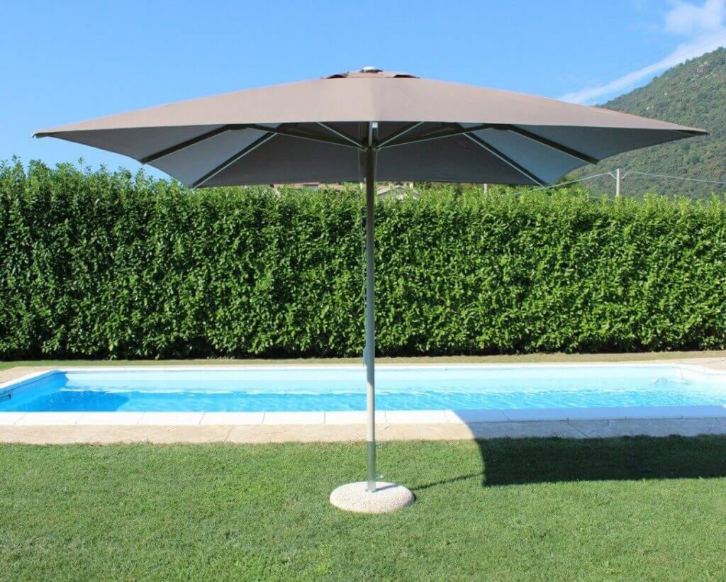 Prospettiva laterale dell'ombrellone Brian a palo centrale posizionato sul prato vicino a una piscina, con siepe sullo sfondo.