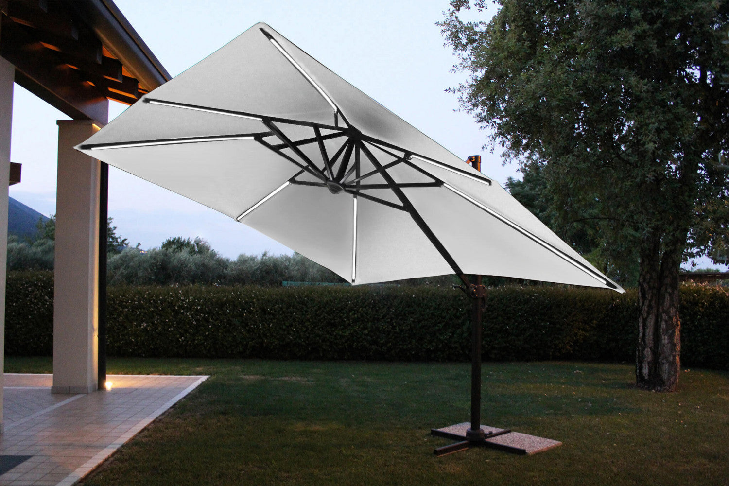 Ombrellone Pitagora con LED integrati nel telaio, aperto in un giardino serale con prato curato e siepe.