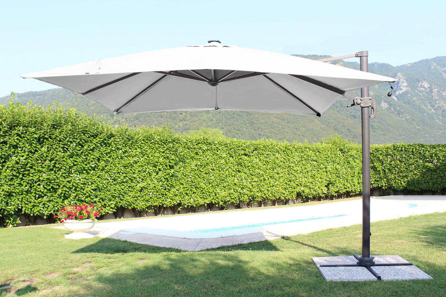 Ombrellone a palo laterale Pitagora aperto di giorno, con telo in poliestere grigio, a bordo di una piscina circondata dal verde.