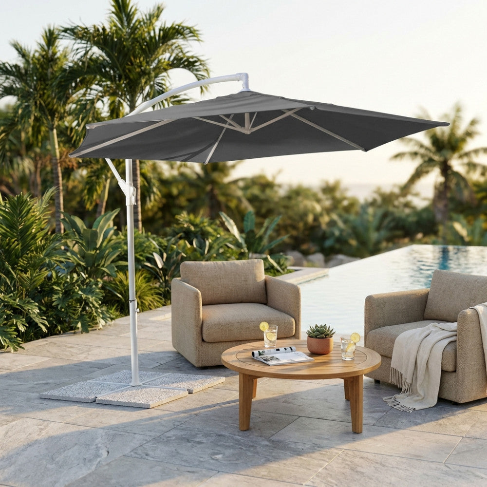 Ombrellone Eris con struttura in acciaio e copertura grigia, posizionato in un giardino tropicale per ombreggiare un salottino lounge con poltrone in tessuto e tavolino in legno.