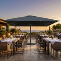 Ombrellone Giove antracite 5x5 aperto su una terrazza panoramica al tramonto con tavoli da pranzo.