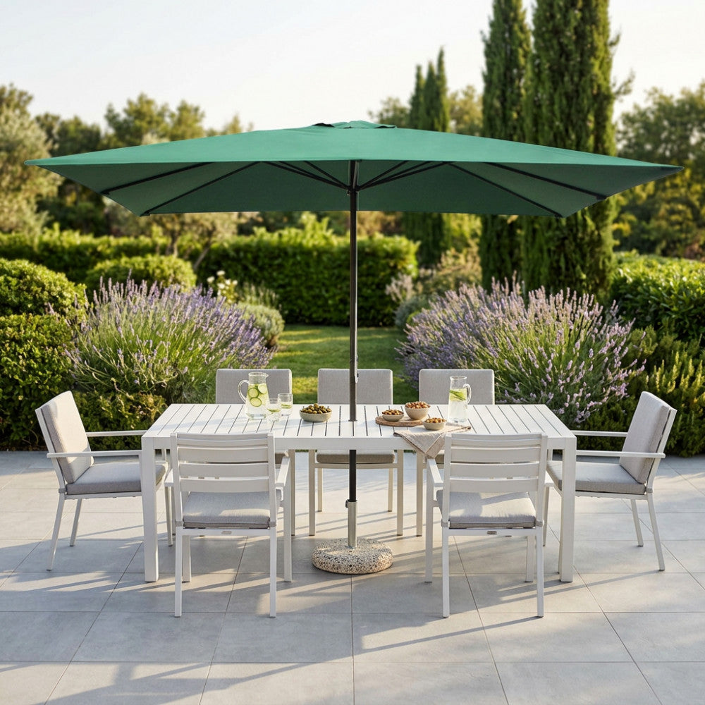 Ombrellone rettangolare Mercurio verde che ombreggia un tavolo da pranzo in metallo bianco in un giardino fiorito.
