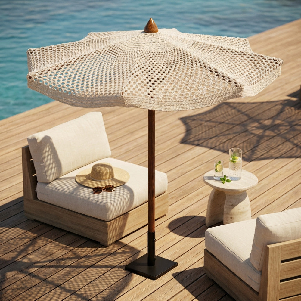 Ombrellone Nador a palo centrale con copertura in cotone macramè beige, posizionato su un ponte in legno a bordo piscina accanto a sedute lounge.