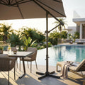 Ombrellone Nettuno Lux aperto a bordo piscina, con struttura in alluminio nero e telo taupe, che ombreggia un tavolo da pranzo elegante.