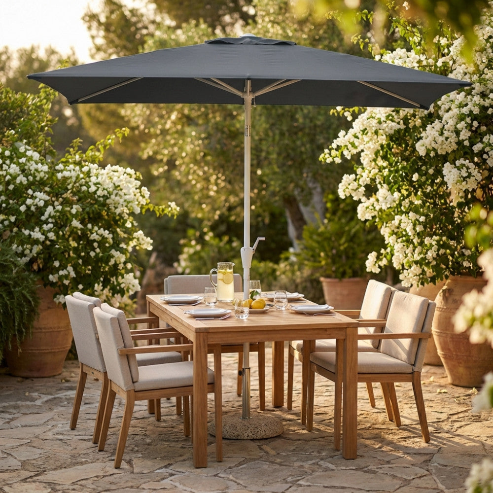 Ombrellone Poly Eco grigio aperto su un tavolo da pranzo in teak in un rigoglioso giardino mediterraneo.