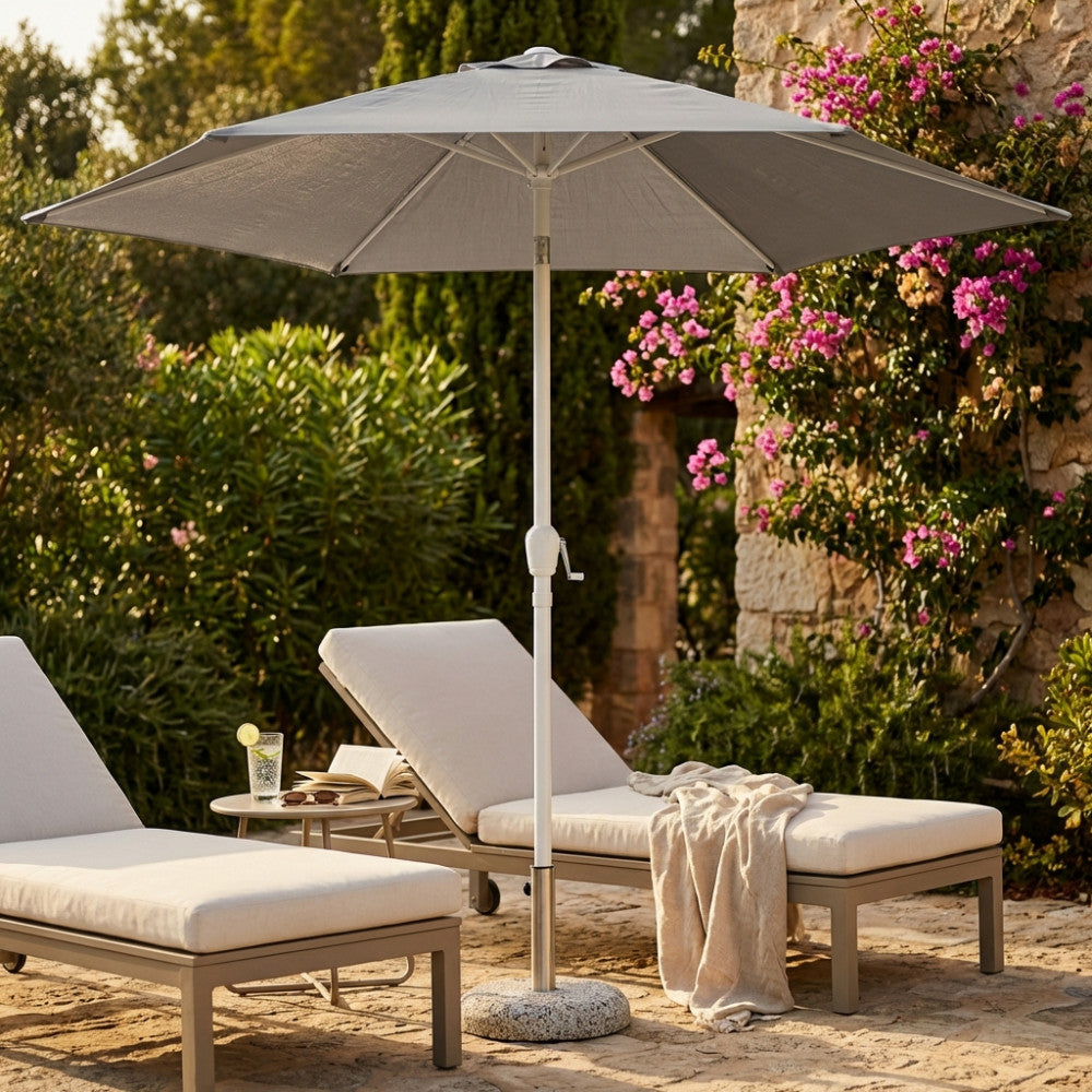 Ombrellone a palo centrale in acciaio e poliestere grigio aperto, posizionato tra due eleganti lettini prendisole in un giardino mediterraneo.